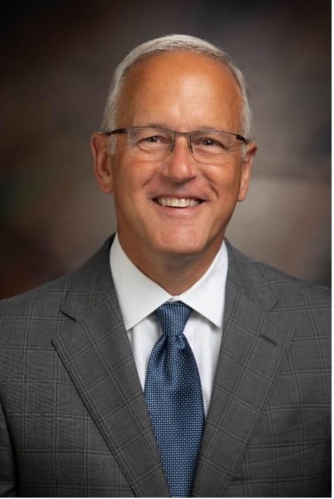 Smiling man with glasses, grey hair, in a gray suit and blue tie.