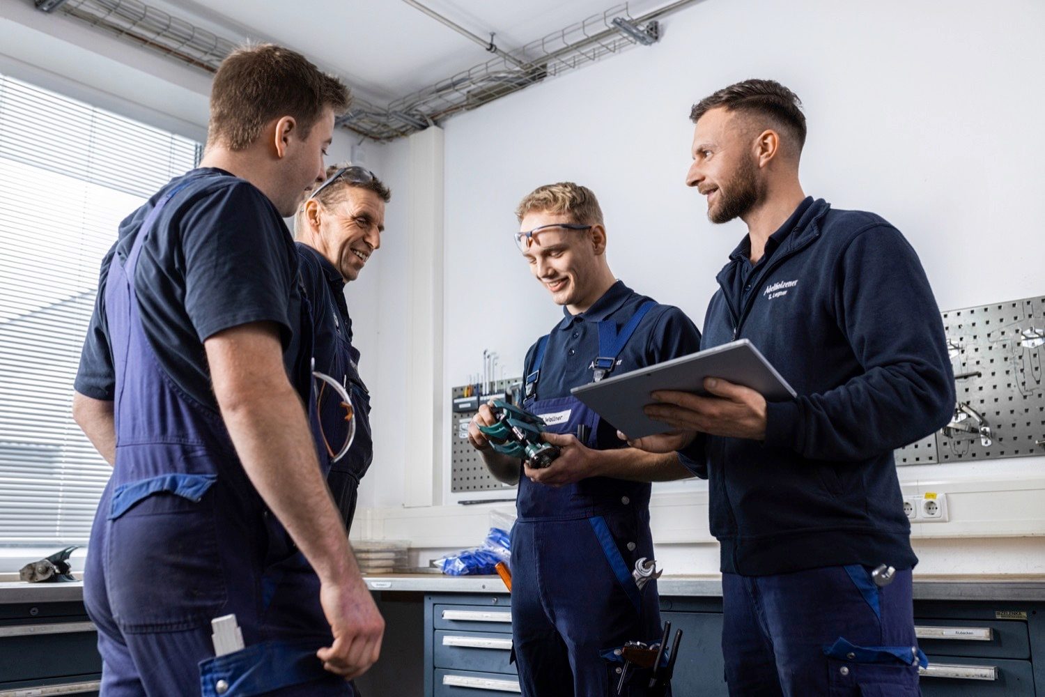 Four men in blue overalls in a workshop, discussing a tool. One holds a tablet.
