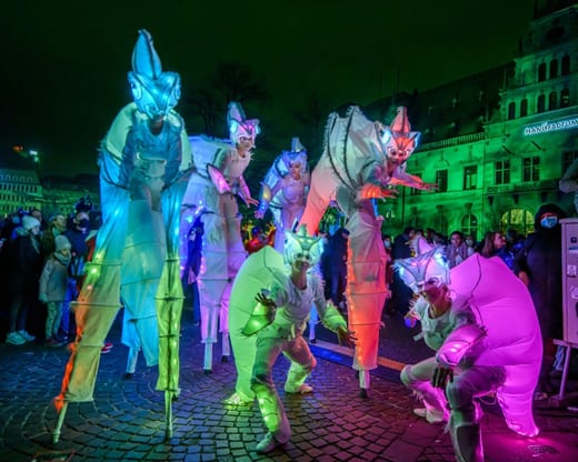 Performers in brightly lit, insect-like costumes on stilts and crouching at a night festival.