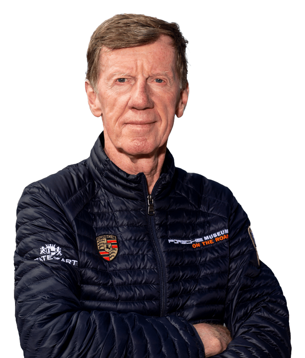 An older man in a blue puffer jacket with Porsche logos, arms crossed.