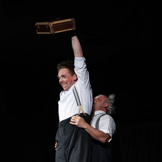 Two smiling clowns on stage; one stands on the other, raising a briefcase in triumph.
