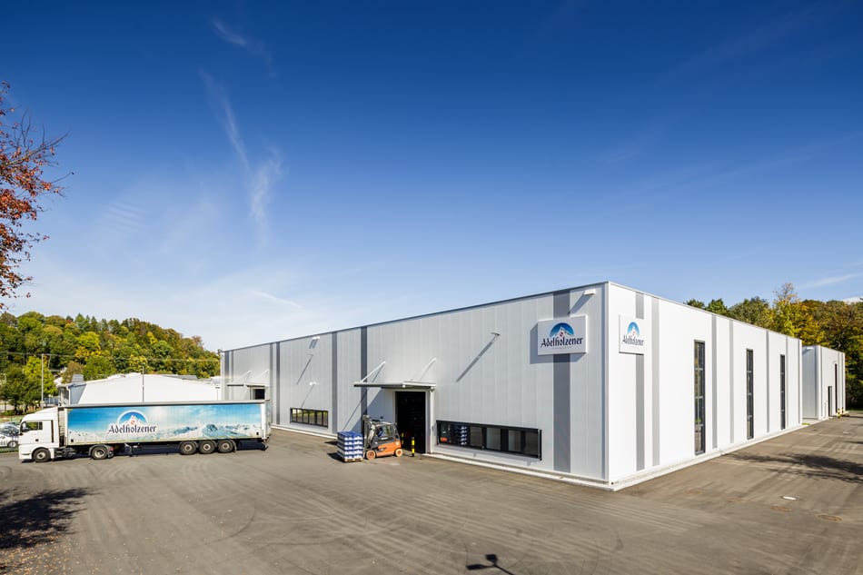 Modern Adelholzener factory with a parked truck and forklift under a clear blue sky.