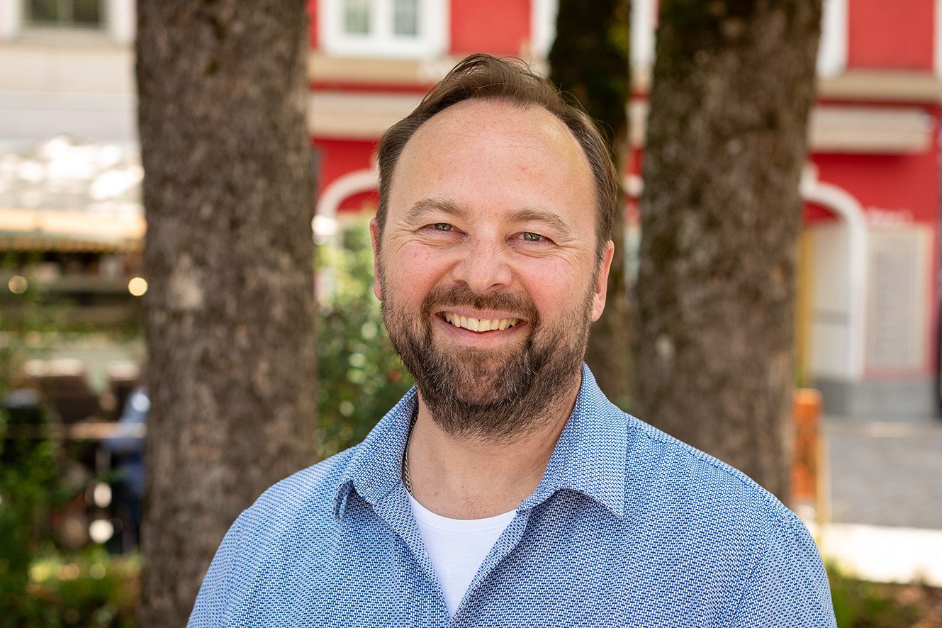 Smiling bearded man in a blue patterned shirt, standing outdoors with trees and a red building in the background.