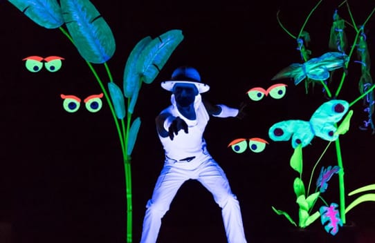 A person in glowing white clothing performing in a dark blacklight setting with neon-colored plants, butterflies, and glowing eyes.