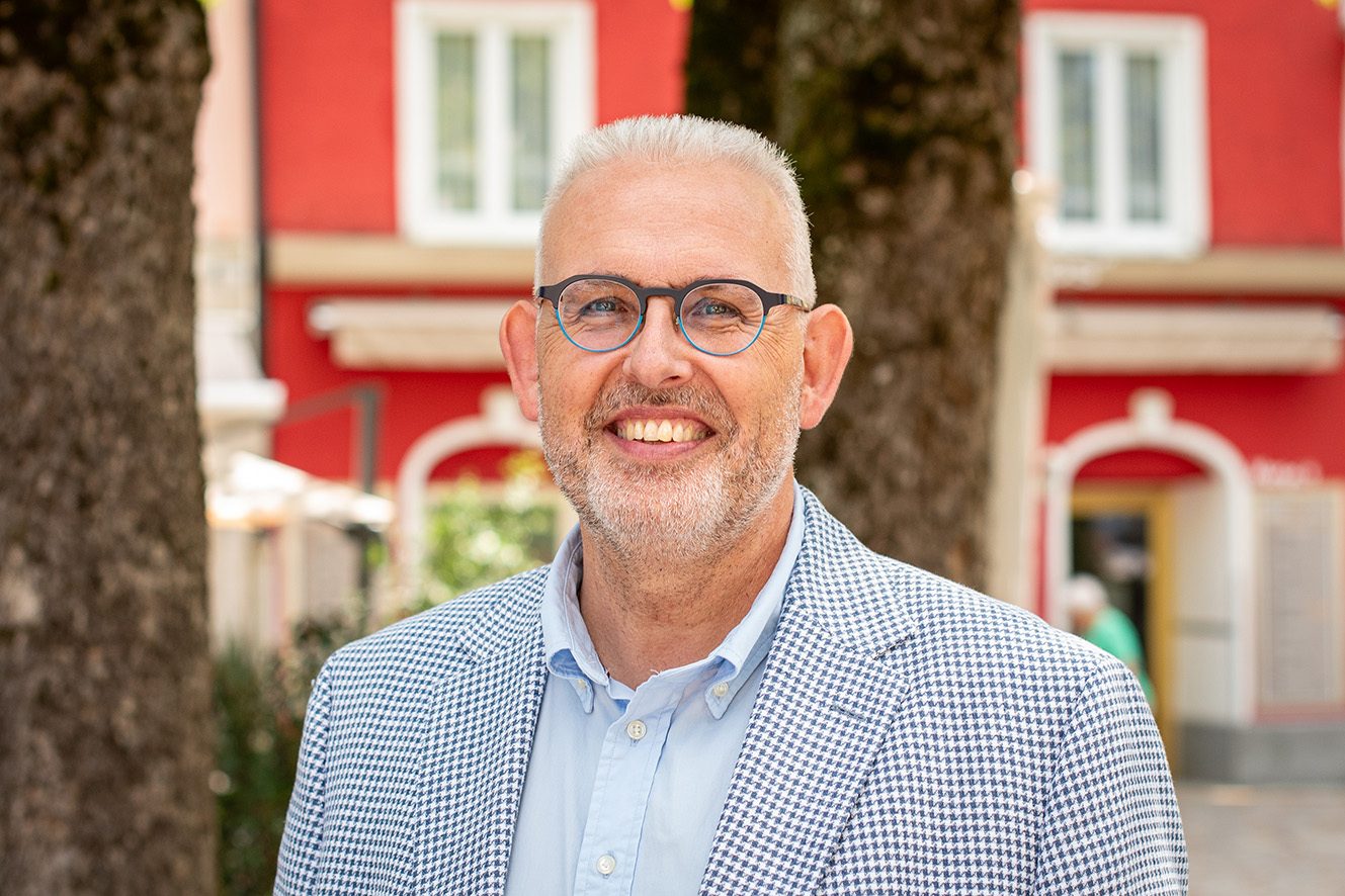 Smiling man with grey hair and beard, wearing glasses and a checkered blazer, standing outdoors.