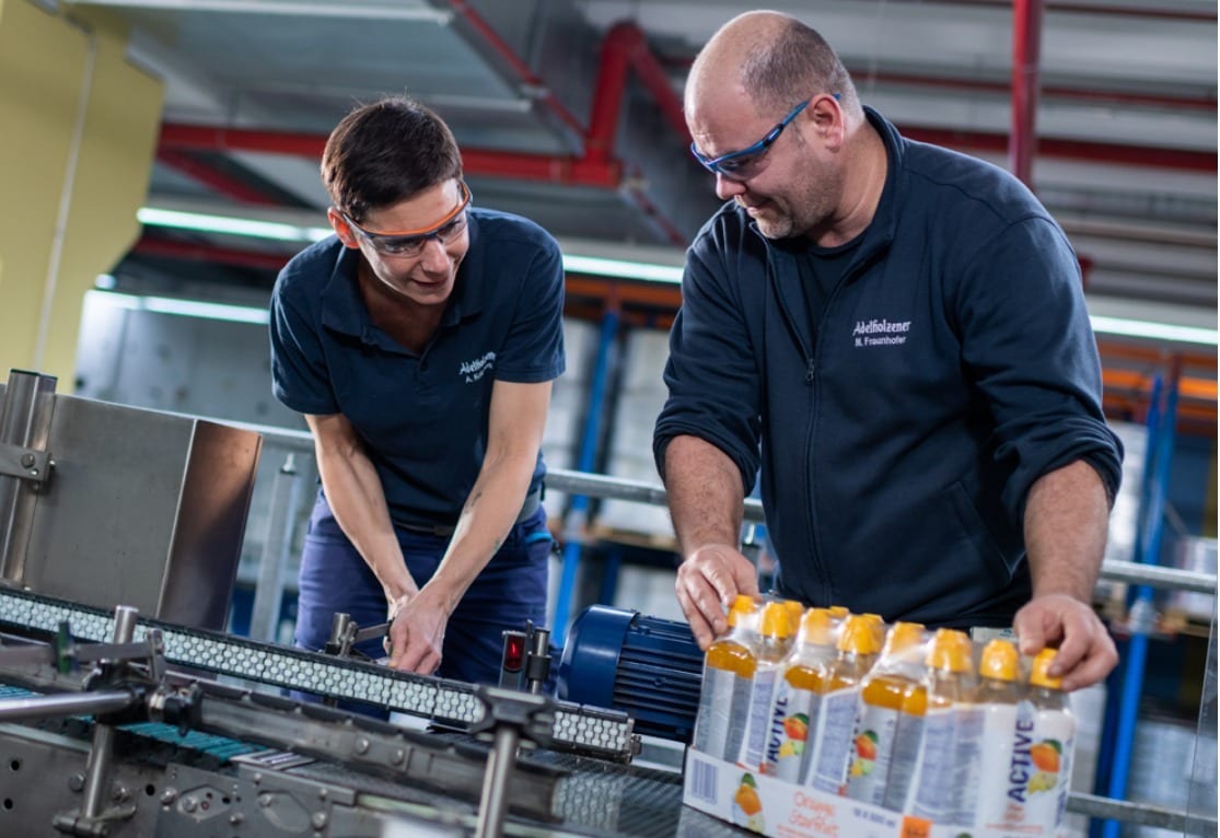 Two factory workers in safety glasses handle juice bottles and equipment.