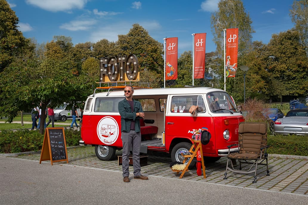 Man stands by a red and white VW photo booth van with a "FOTO" sign, chalkboard, and props.