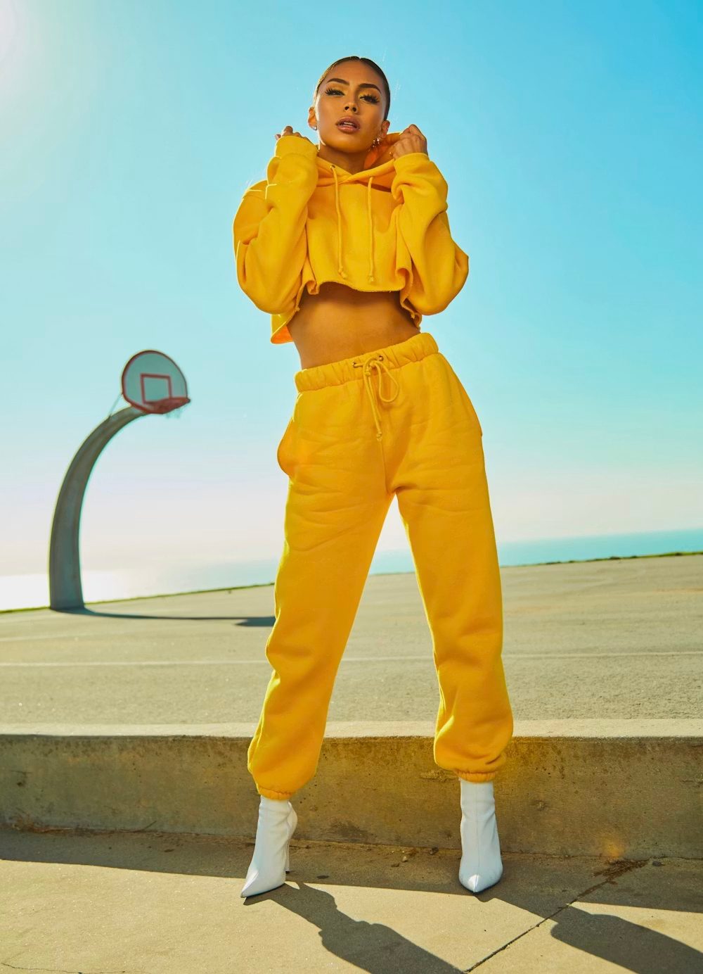 A stylish woman in a yellow cropped hoodie and sweatpants with white boots on an outdoor court.