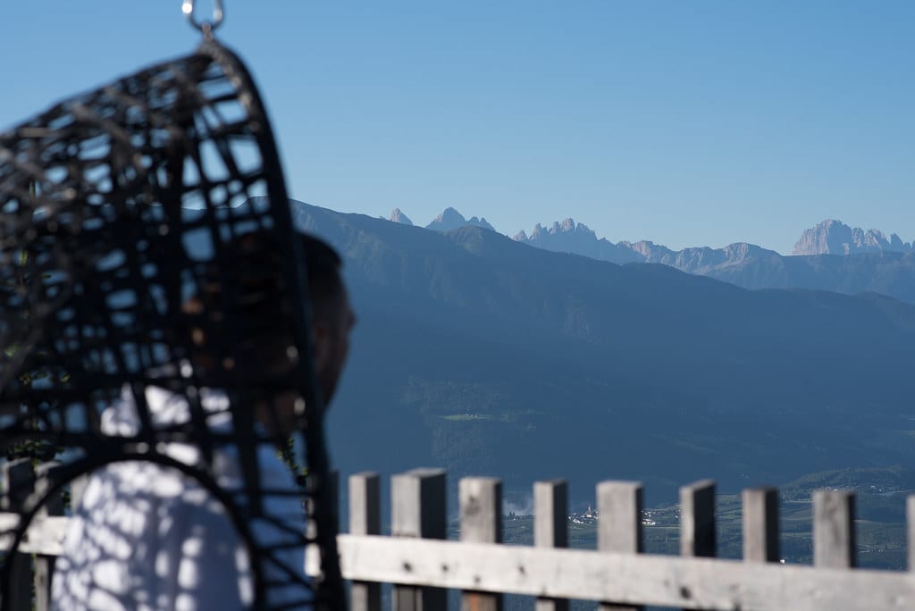 Person in a hanging chair enjoys a panoramic view of a mountain range and valley.