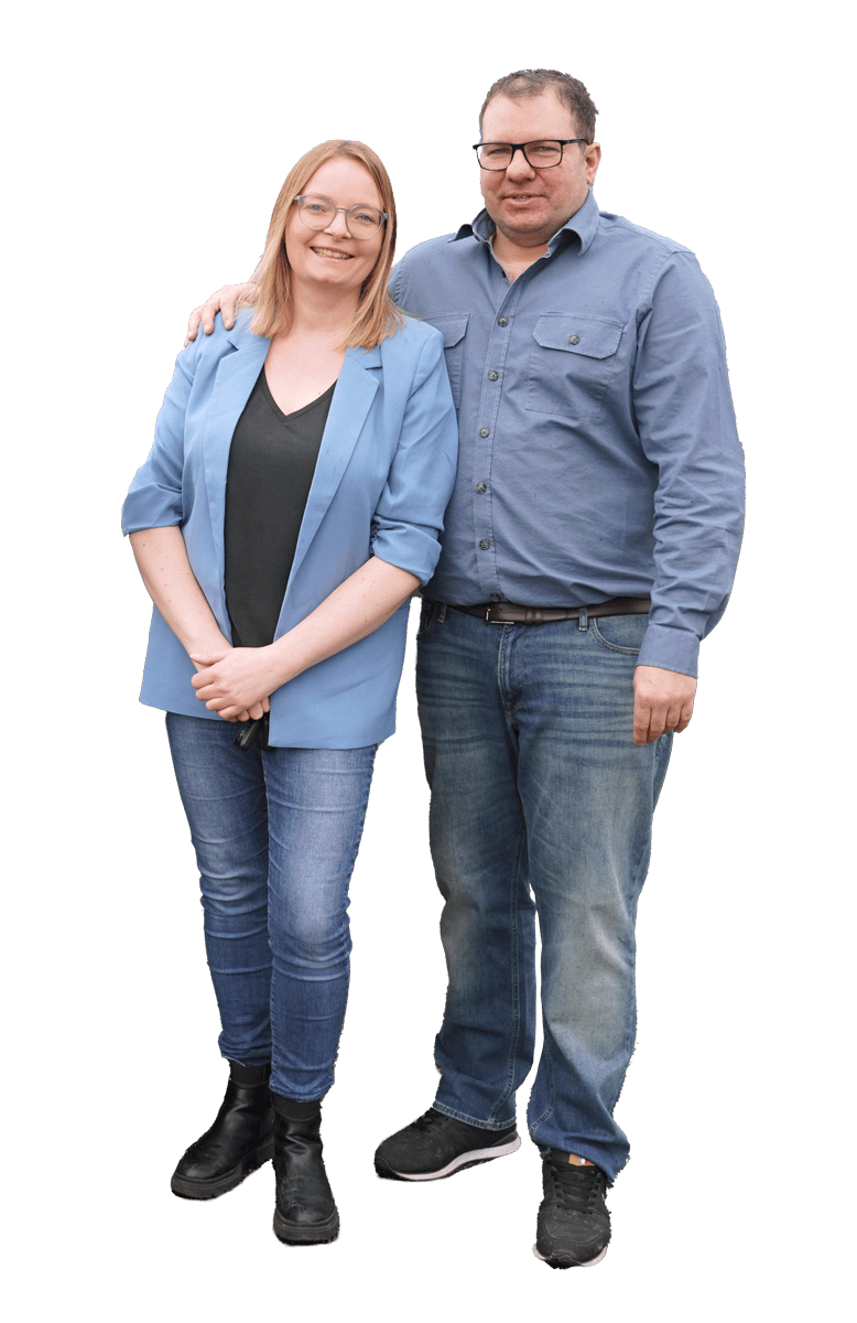 A smiling man and woman, both in jeans and glasses, stand together, arm in arm.