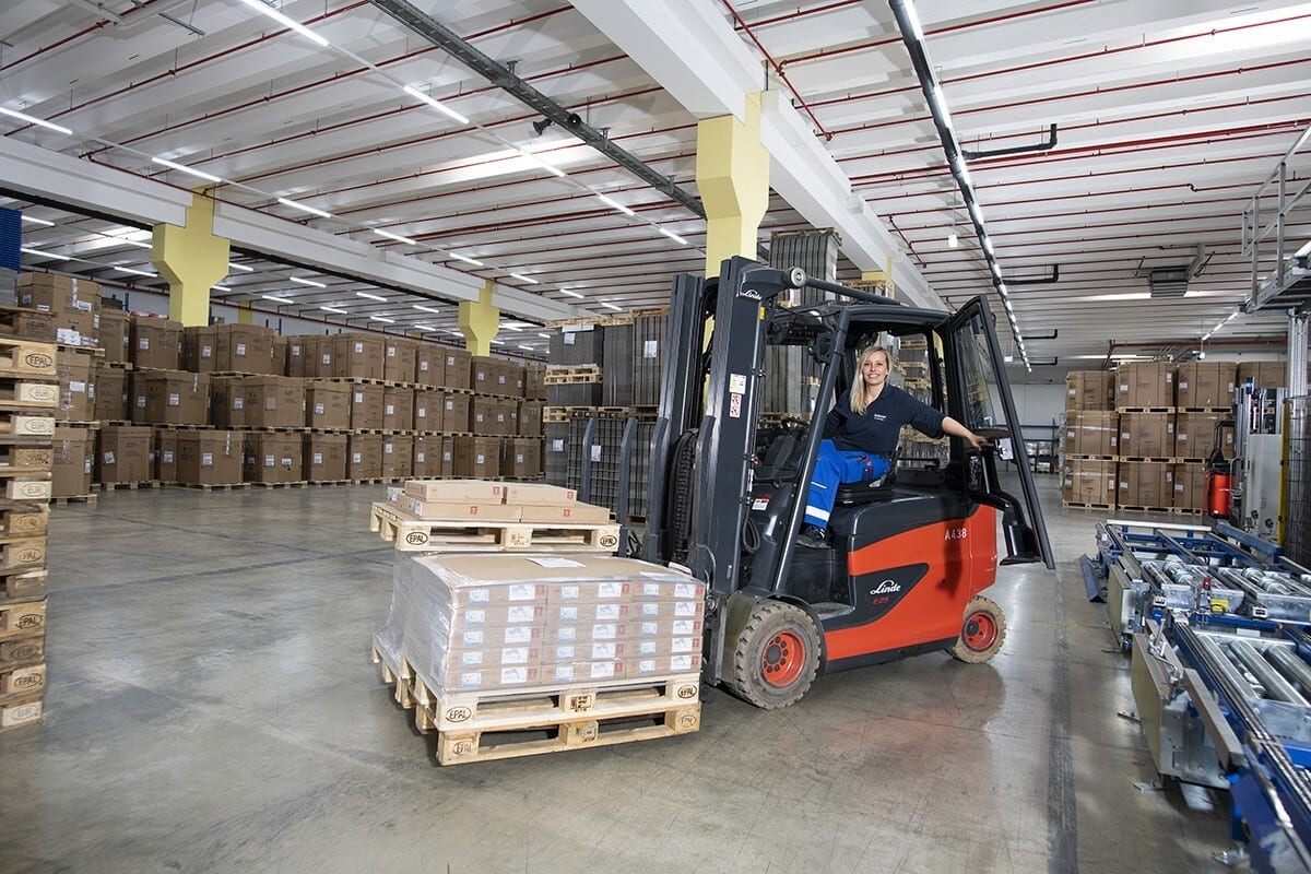 Woman operates a forklift in a warehouse with stacked boxes.