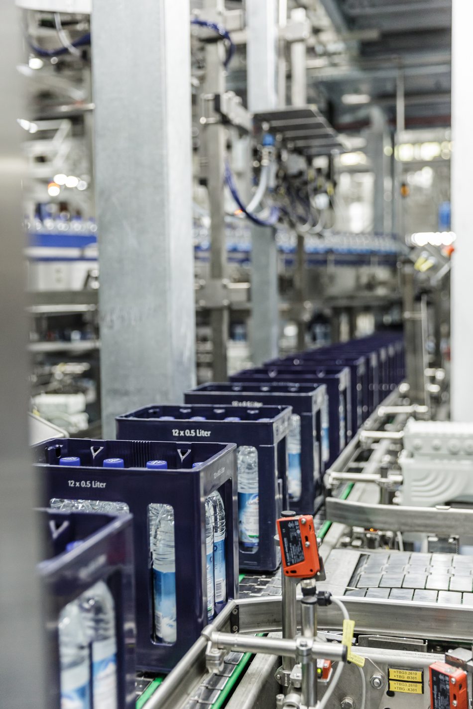 Factory line with blue crates of 12 x 0.5 liter water bottles on a conveyor belt.