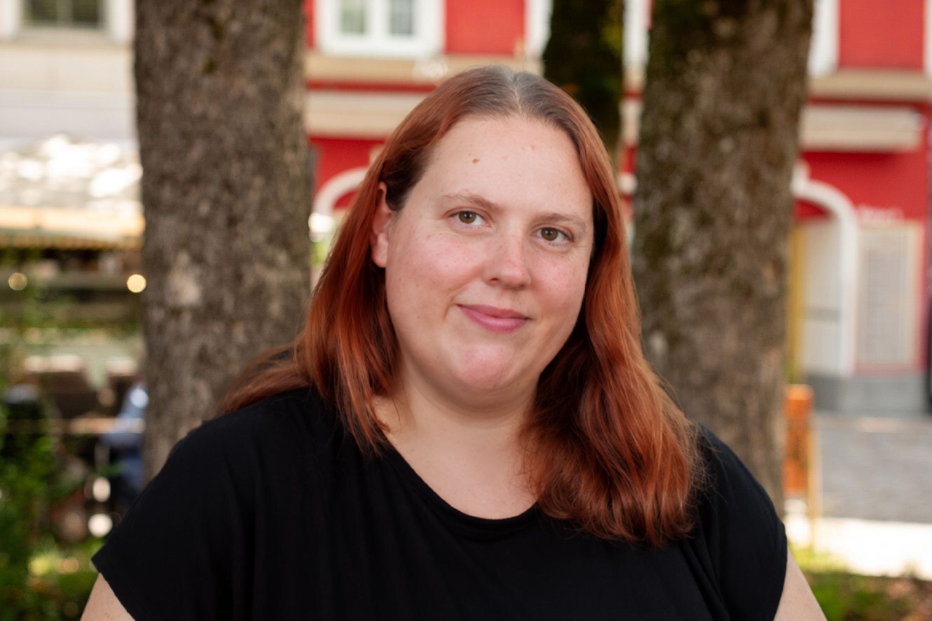 A smiling woman with reddish-brown hair and a black top, looking at the camera outdoors.