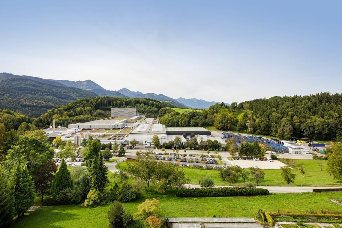 Industrial complex in a green valley, surrounded by mountains and forests under a clear sky.