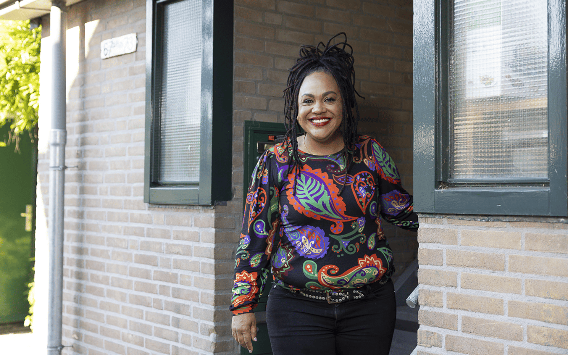 Smiling Black woman with dreadlocks in a colorful shirt stands in a doorway of a brick building.