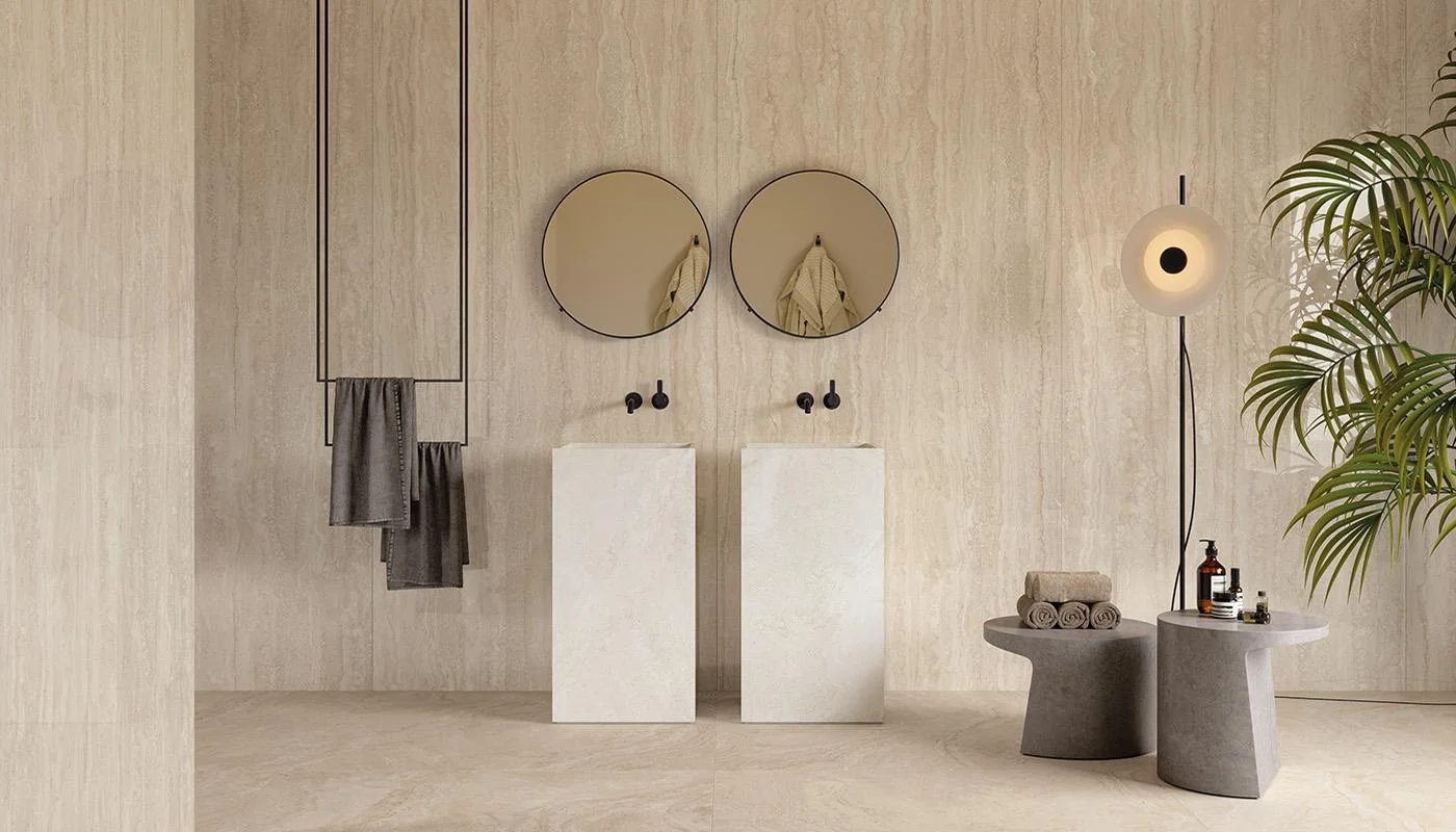 Minimalist bathroom with light tiled walls, twin pedestal sinks, round mirrors, and a floor plant.