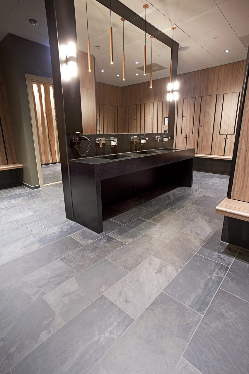 Modern locker room with gray tiled floor, dark sinks, wood lockers, and mirror.