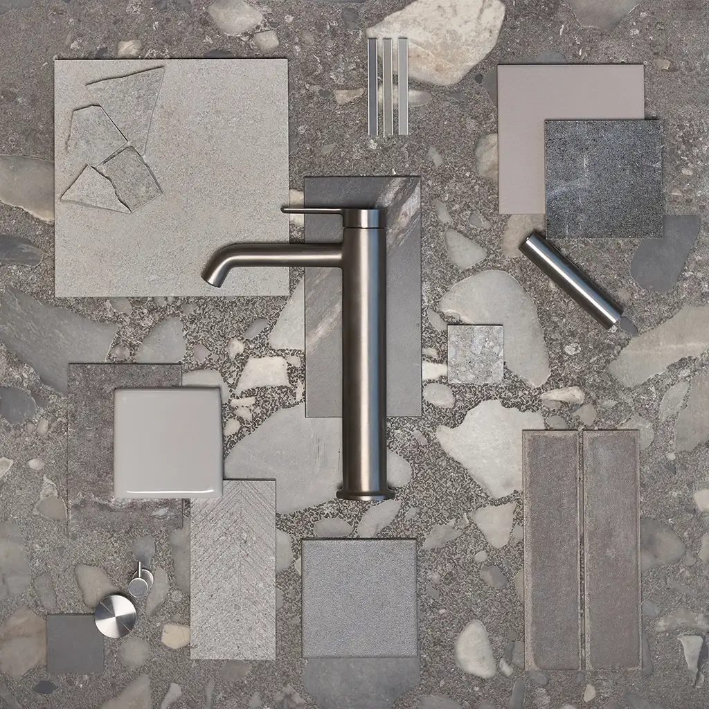 A flat lay of various grey and white tiles, stone samples, and a brushed metal faucet on a terrazzo background.