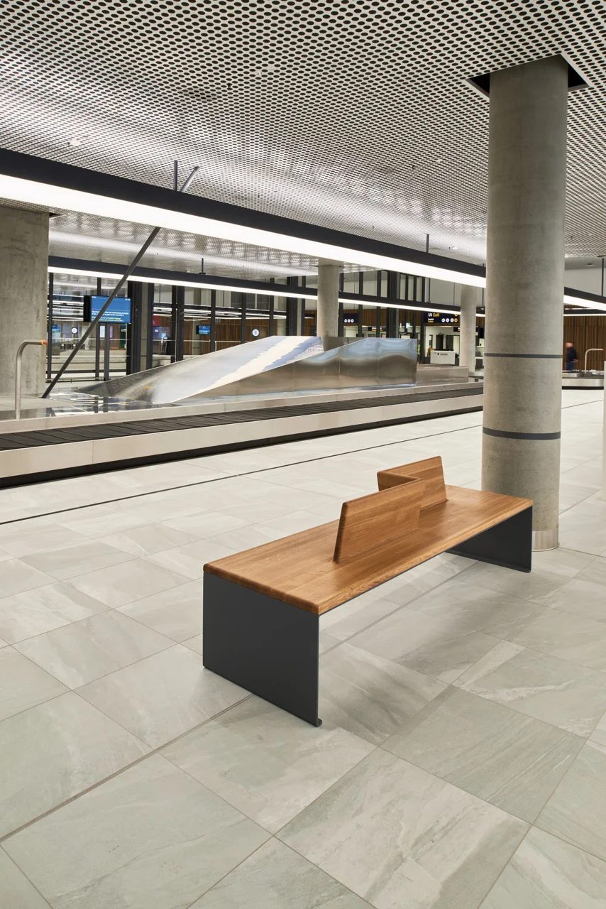 Modern airport baggage claim with wooden bench, baggage carousel, and perforated ceiling.