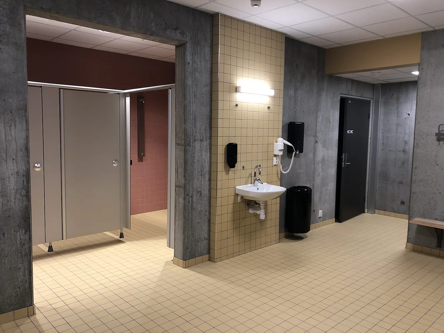 Modern public restroom with concrete walls, light tiled floor, toilet stalls, and a sink area.