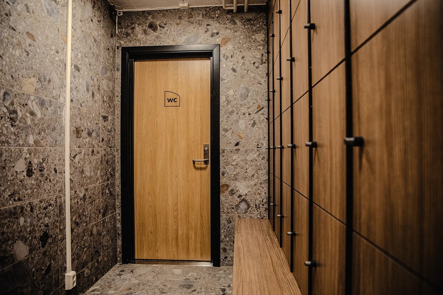A modern corridor with a wooden WC door, speckled tiled walls, wooden lockers, and a bench.