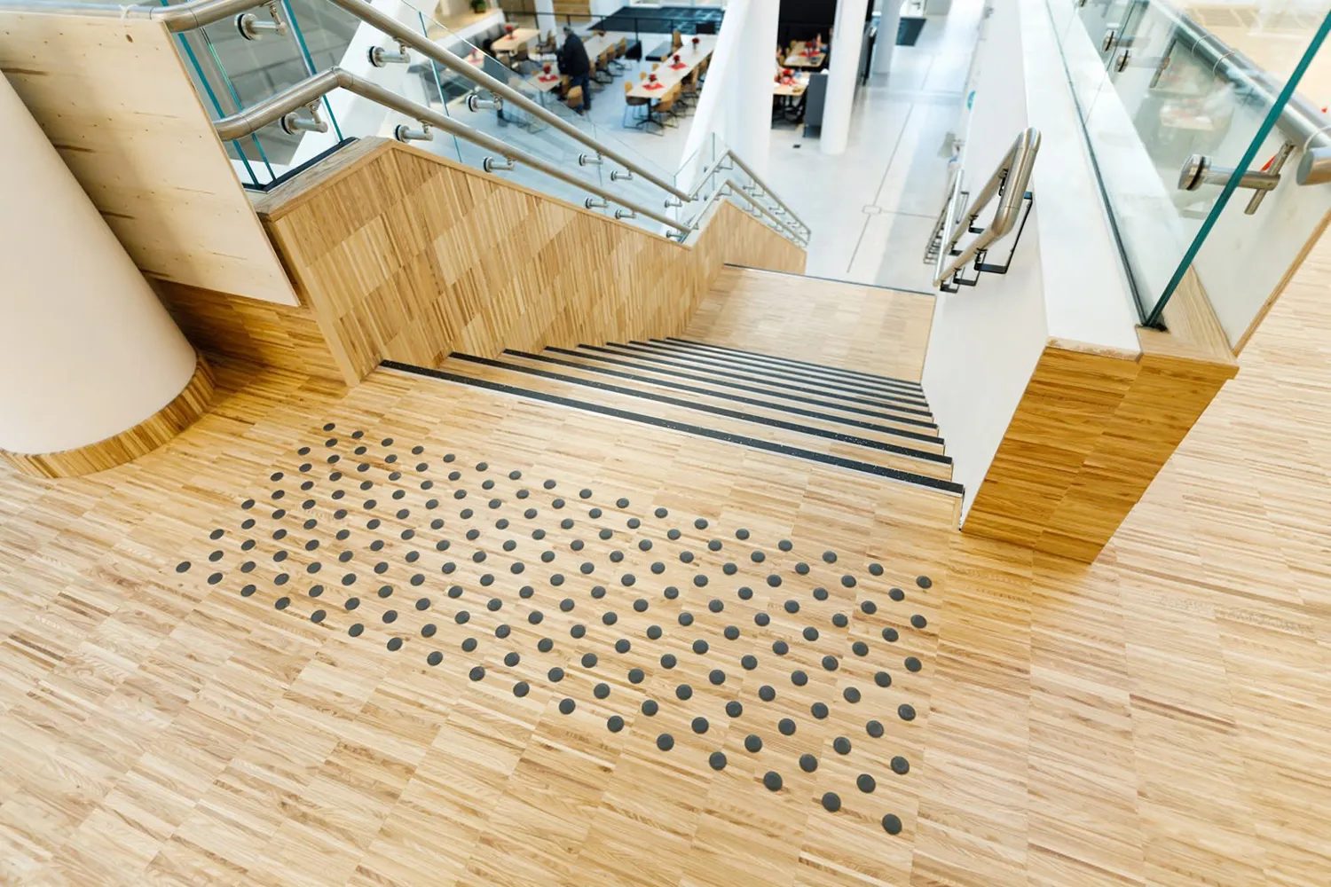Downward view of light wooden stairs with black tactile dots and glass railings.