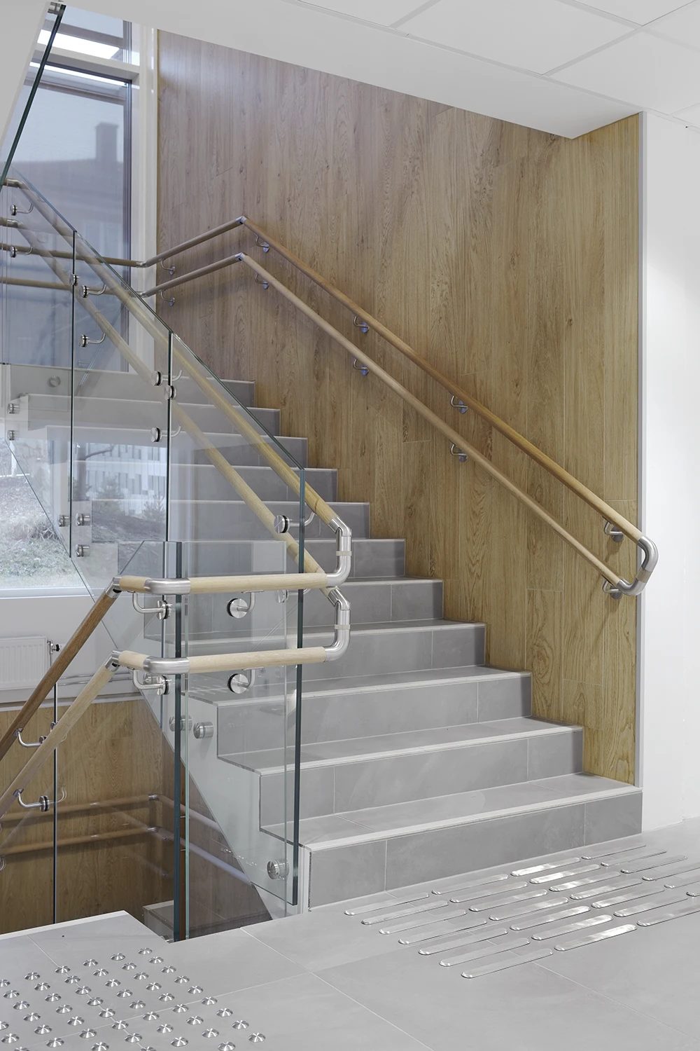 Modern staircase with glass and wood railings, grey steps, wood wall, and tactile flooring.
