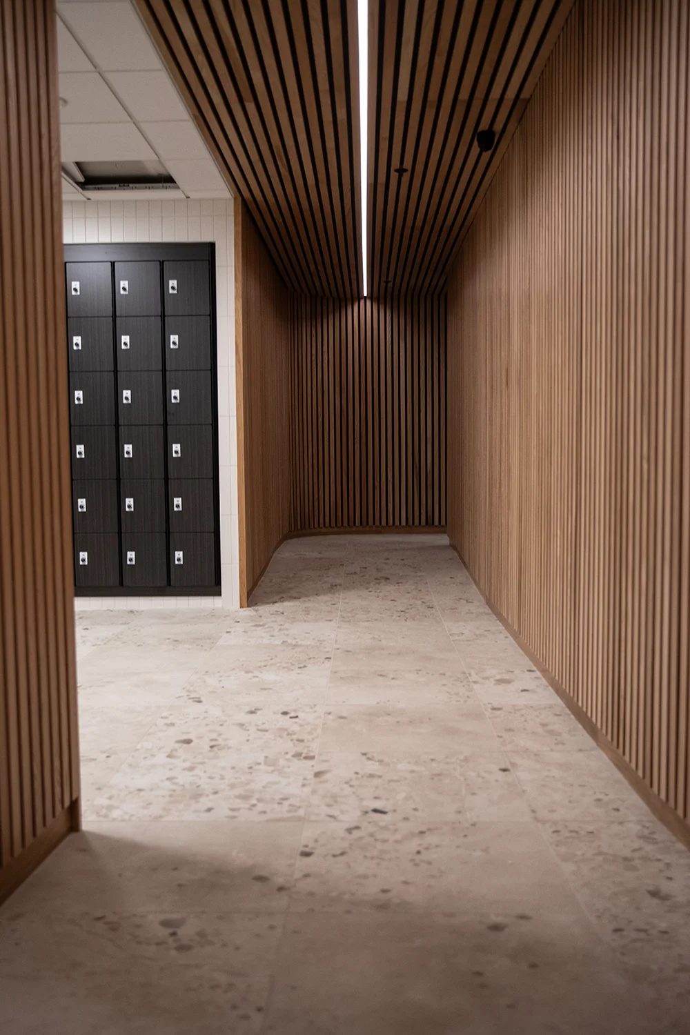 Modern hallway with wood slat walls and ceiling, dark lockers, and speckled floor tiles.
