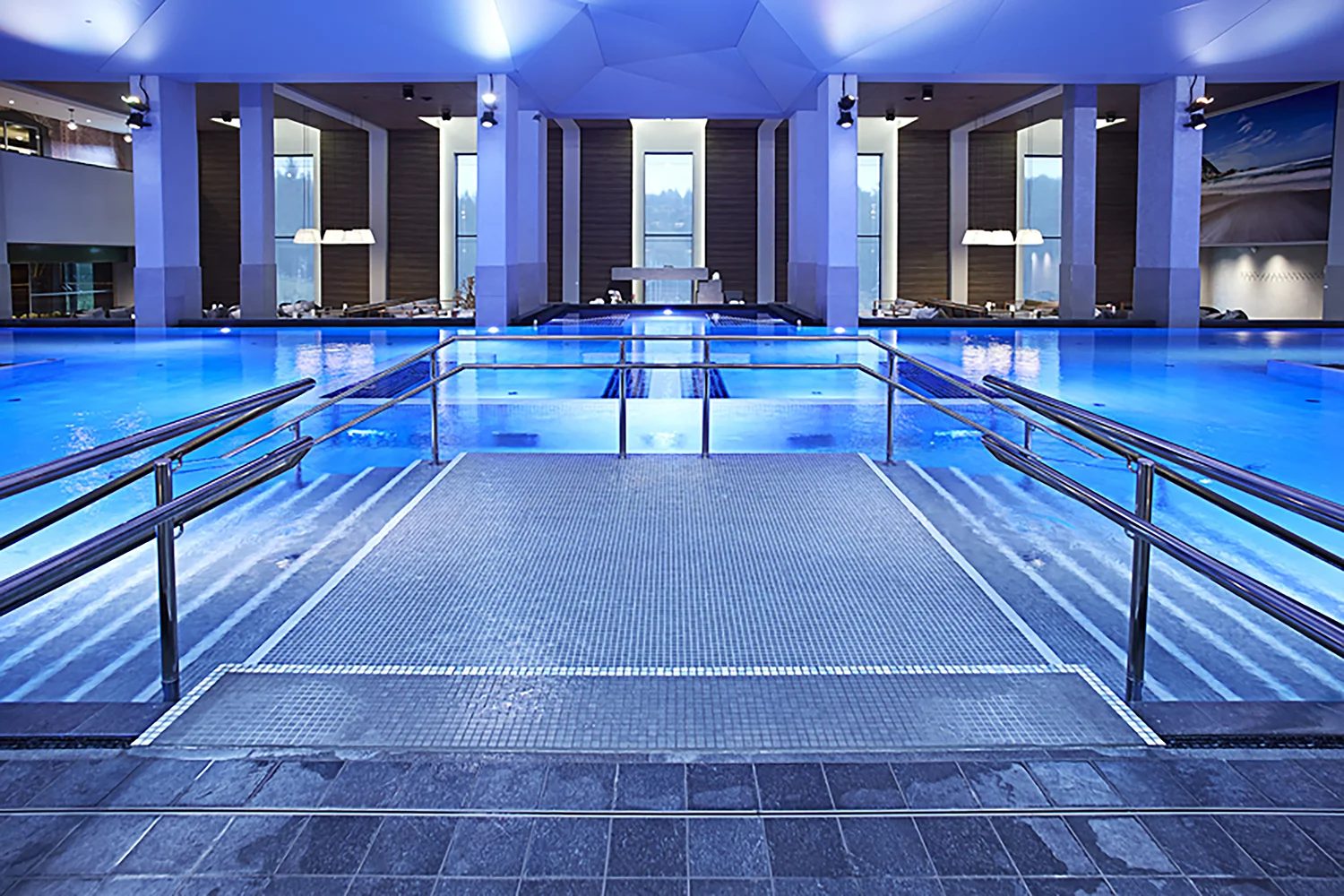 A modern indoor swimming pool with glowing blue water and accessible entry ramps.
