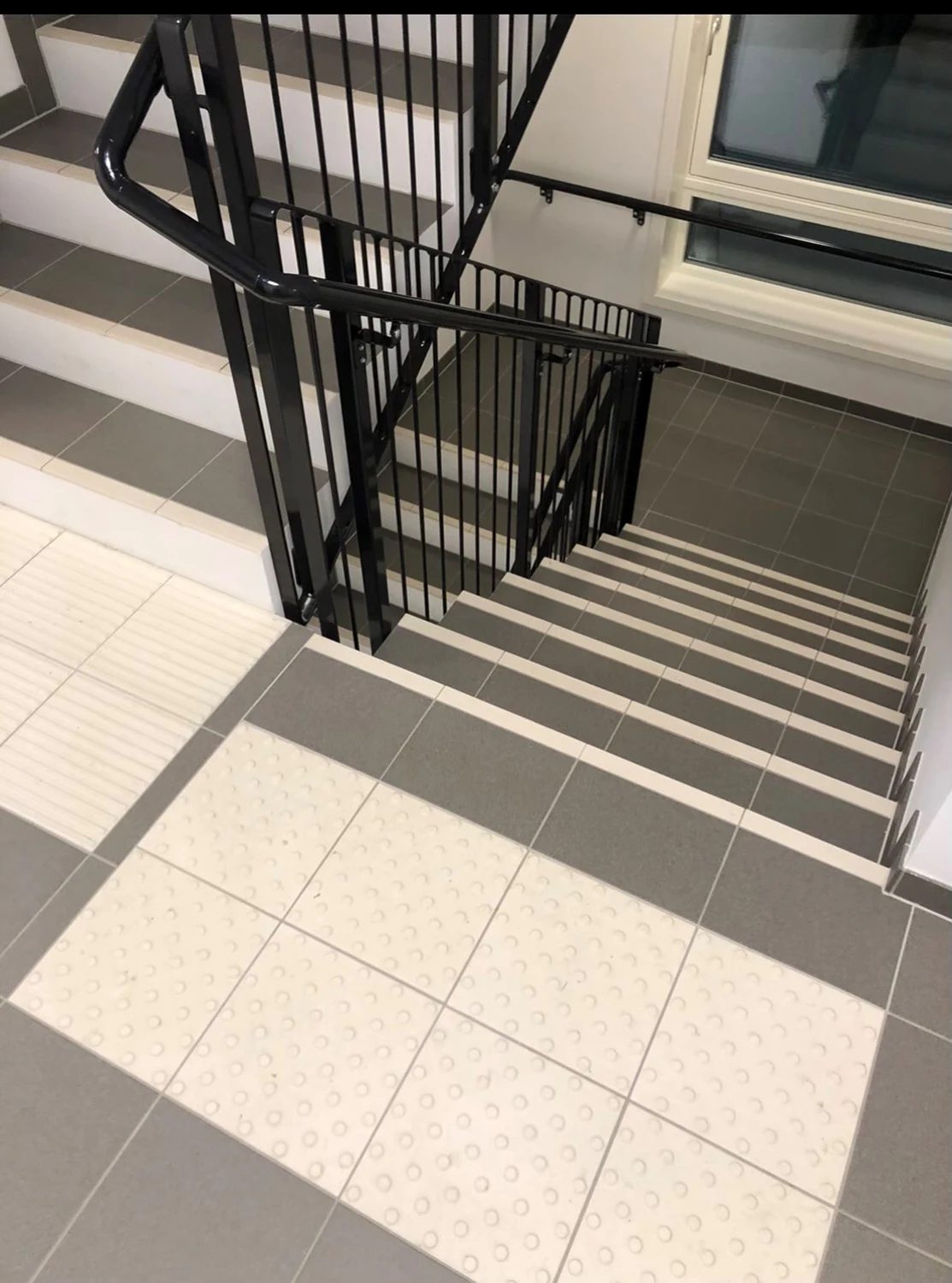Modern staircase with black railing, grey and white tiled steps, tactile floor, and window.