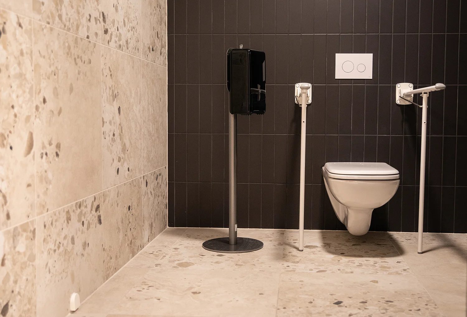 Accessible restroom with wall toilet, grab bars, sanitizer, dark and light speckled tiles.