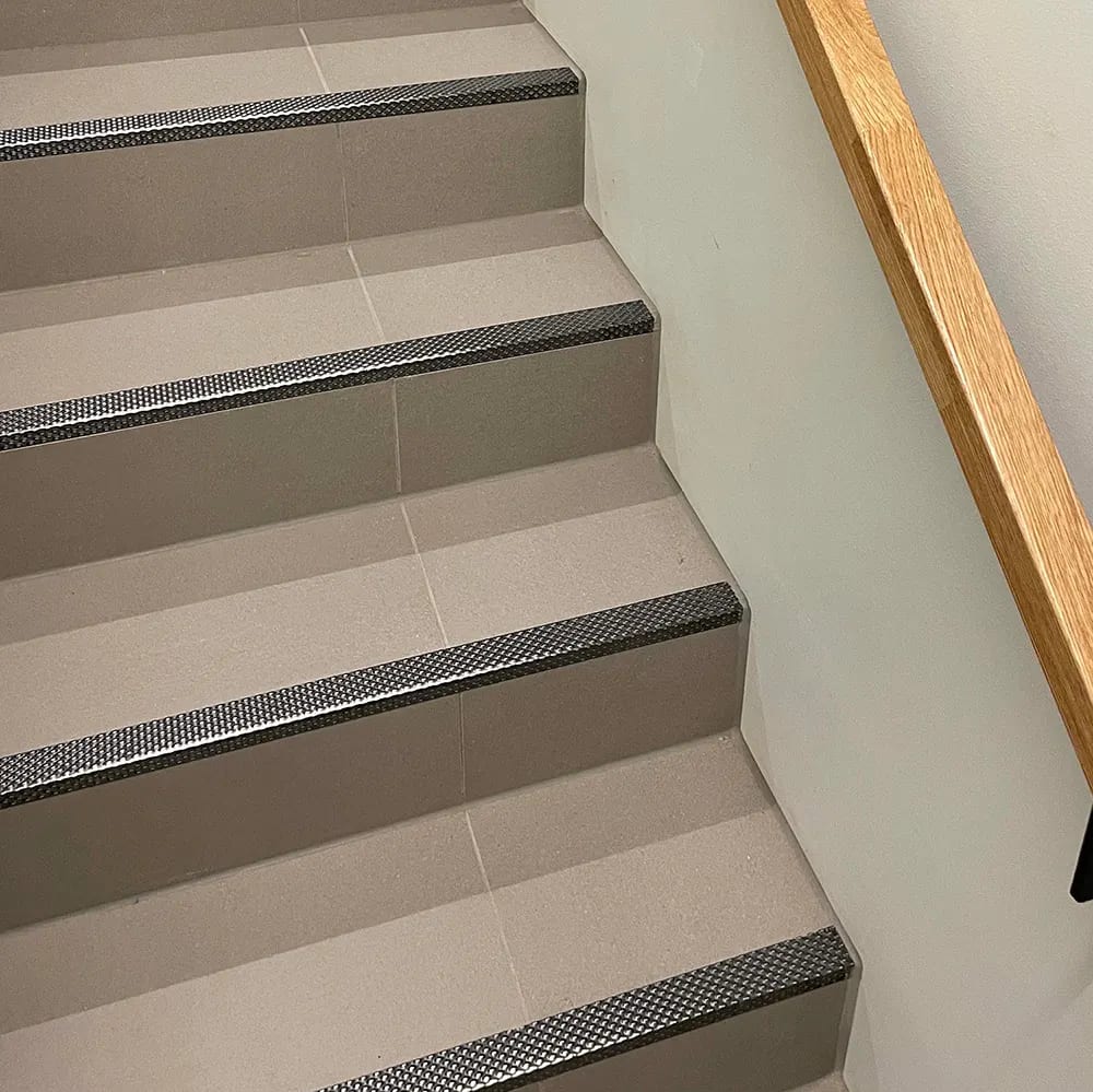 Light-colored tiled stairs with dark, textured anti-slip strips on each step and a wooden handrail.