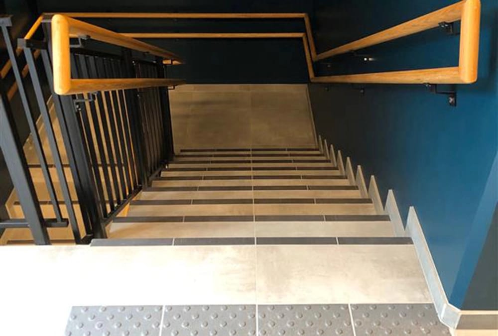Modern stairs descending with wooden handrails, black railing, and dark blue wall.