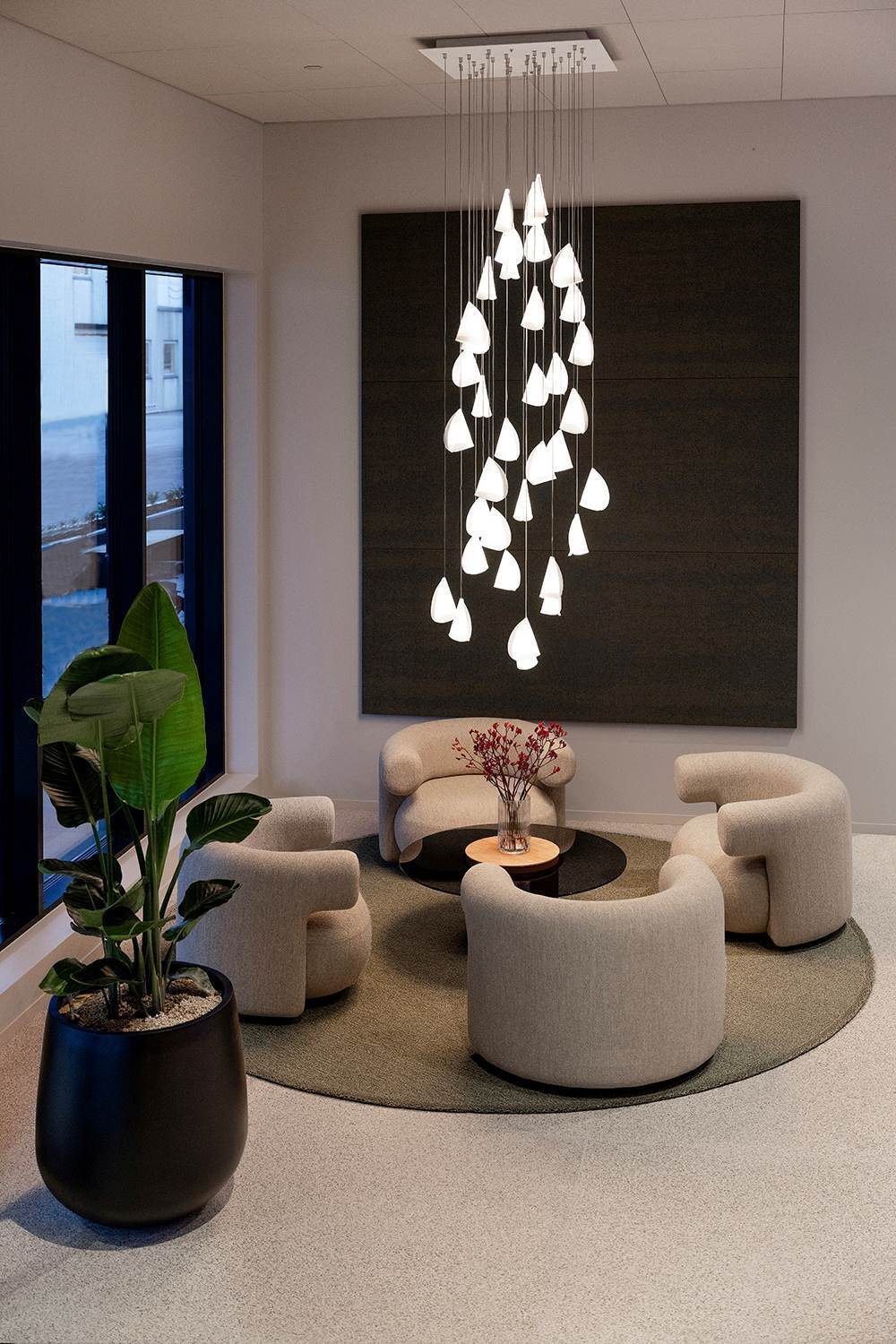 Modern lounge with cascading chandelier, curved chairs, potted plant, and large window.