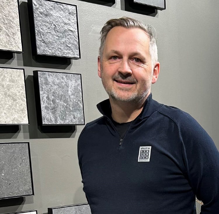 A smiling man in a dark blue sweater stands in front of tile samples.