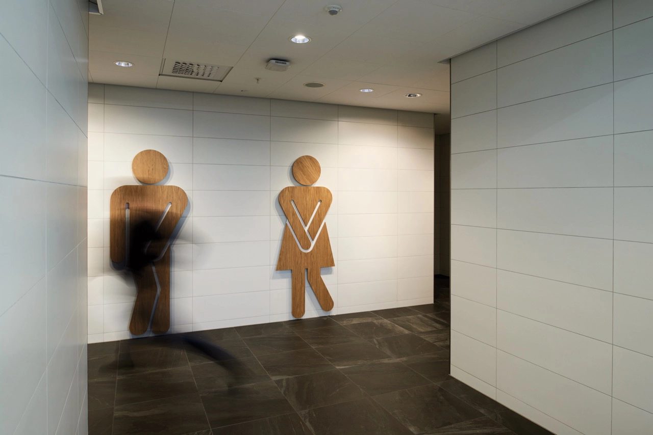 Wooden male and female restroom signs in urgent poses on a white wall, dark floor, with a blurred shadow.