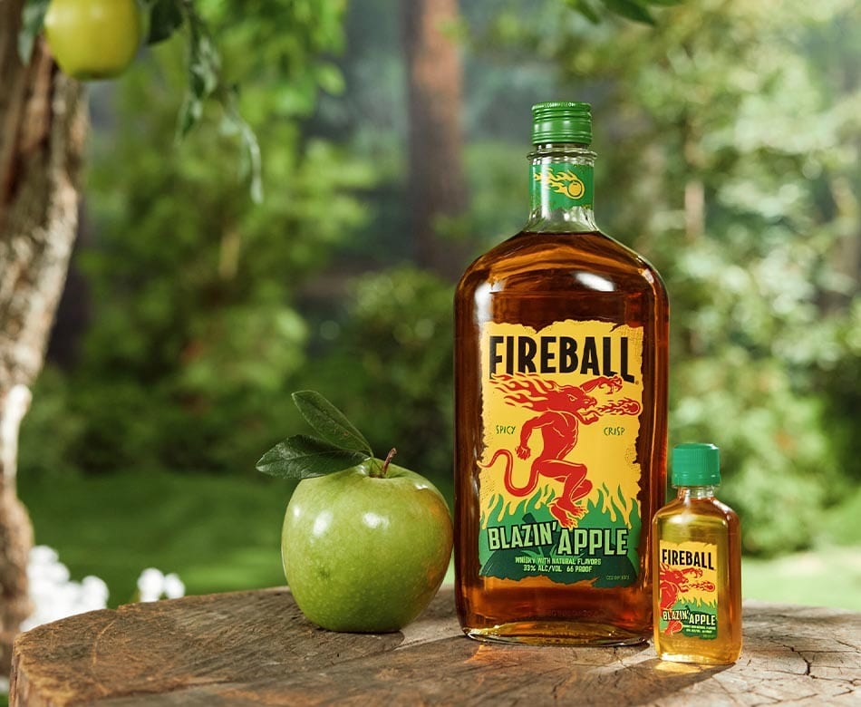 Two Fireball Blazin' Apple whiskey bottles and green apples on a wooden stump outdoors.
