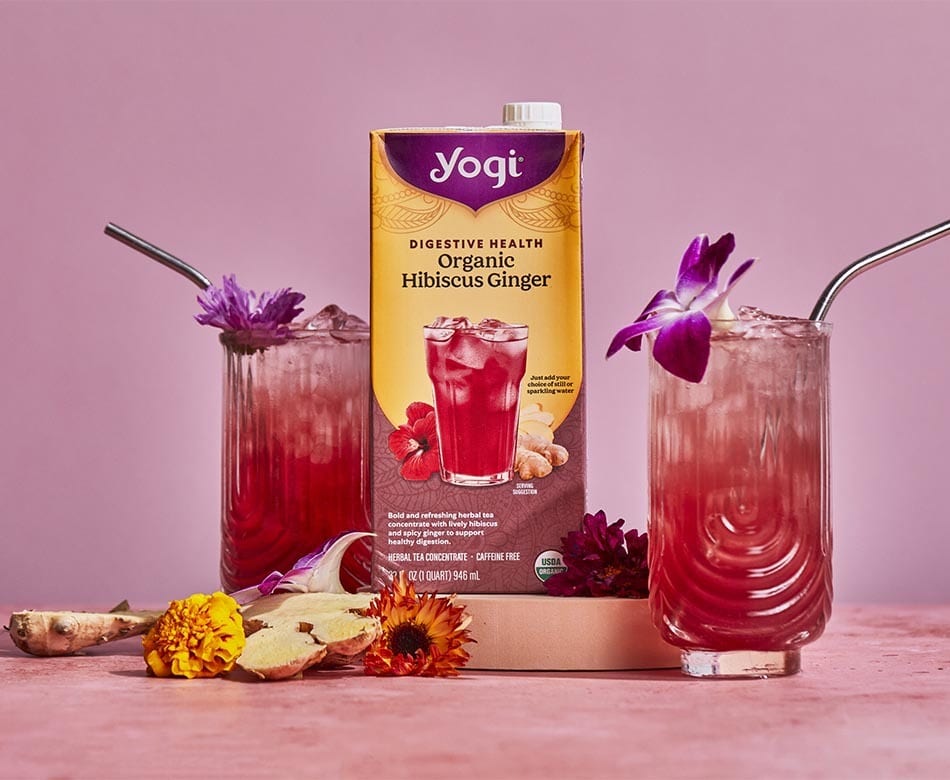 Yogi Hibiscus Ginger tea carton & two iced drinks with flowers and ginger on a pink surface.