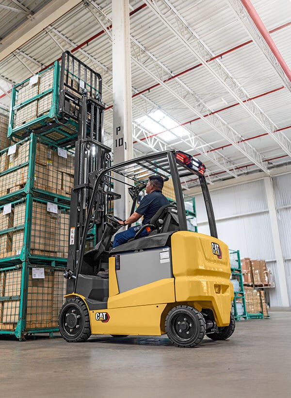 Man on yellow forklift lifting caged pallets in warehouse.