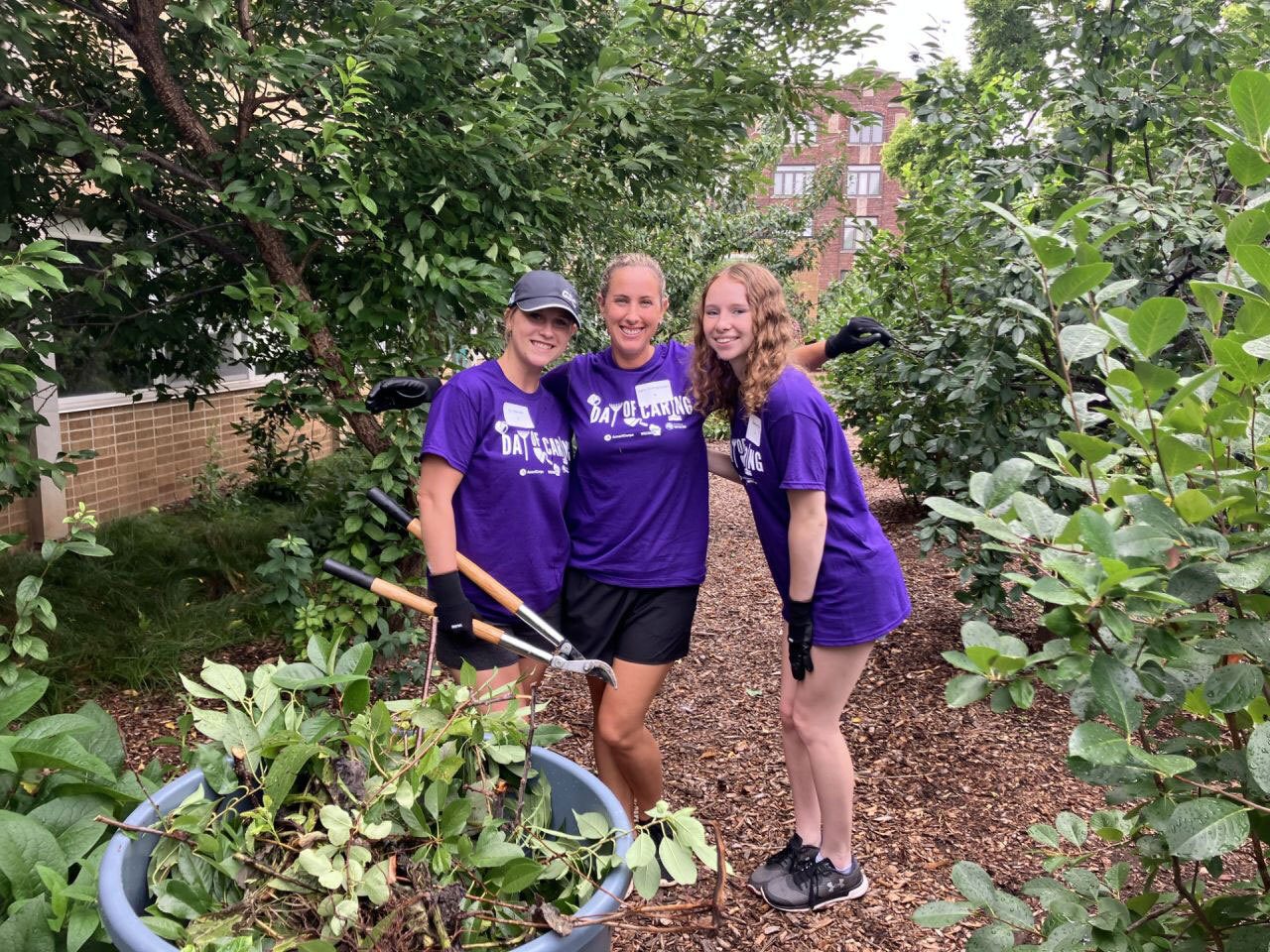 Three women in "Day of Caring" shirts smile while gardening outdoors.