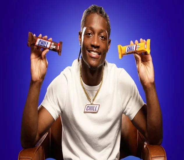 A smiling man in a white t-shirt and gold chain holds up two 'Chill' ice cream bars against a blue background.