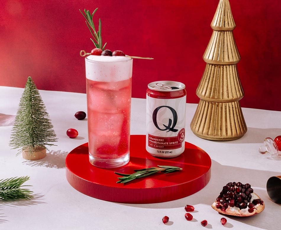 Pink cranberry pomegranate cocktail, Q Spritz can, and Christmas decor on a red and white background.