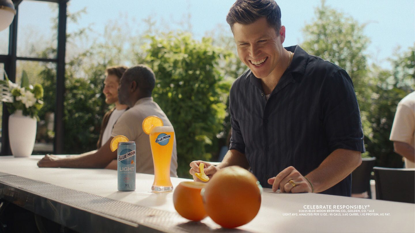 Smiling man peels orange next to Blue Moon beer on an outdoor bar.
