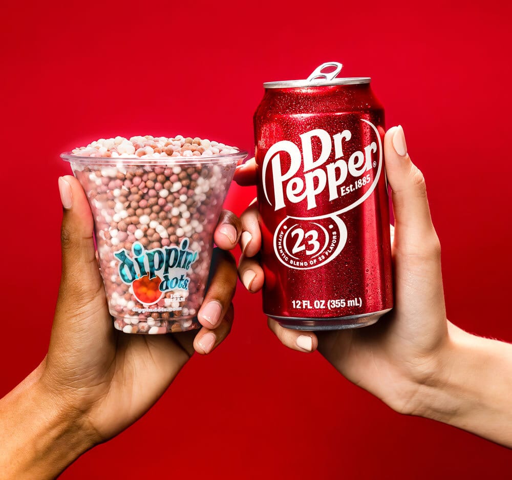 Dippin' Dots cup and Dr Pepper can held by two hands on red background.