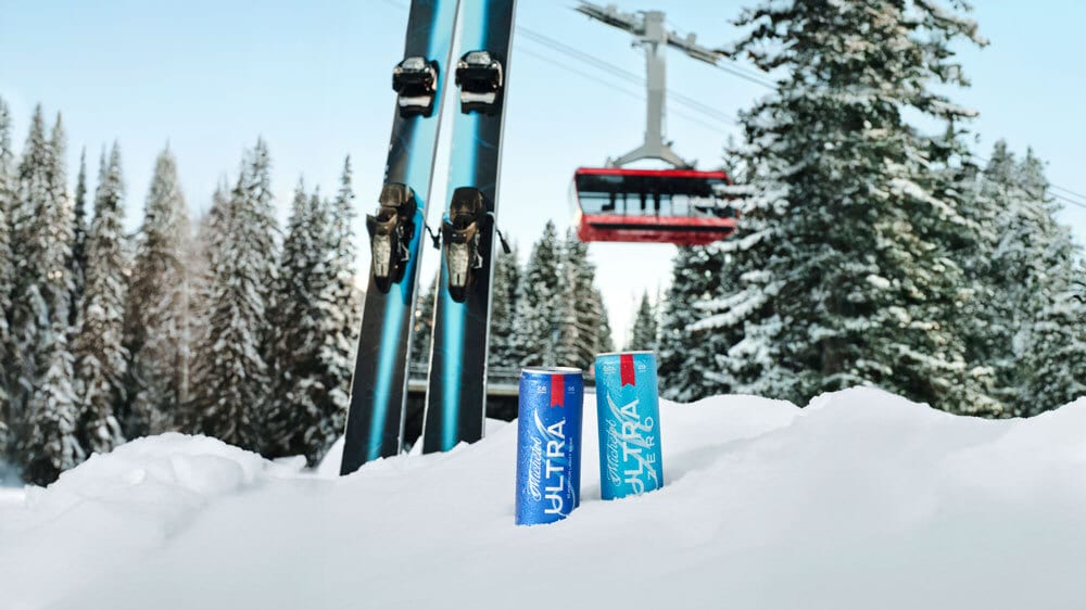 Two Michelob Ultra cans in snow, with skis, pine trees, and a ski lift in the background.