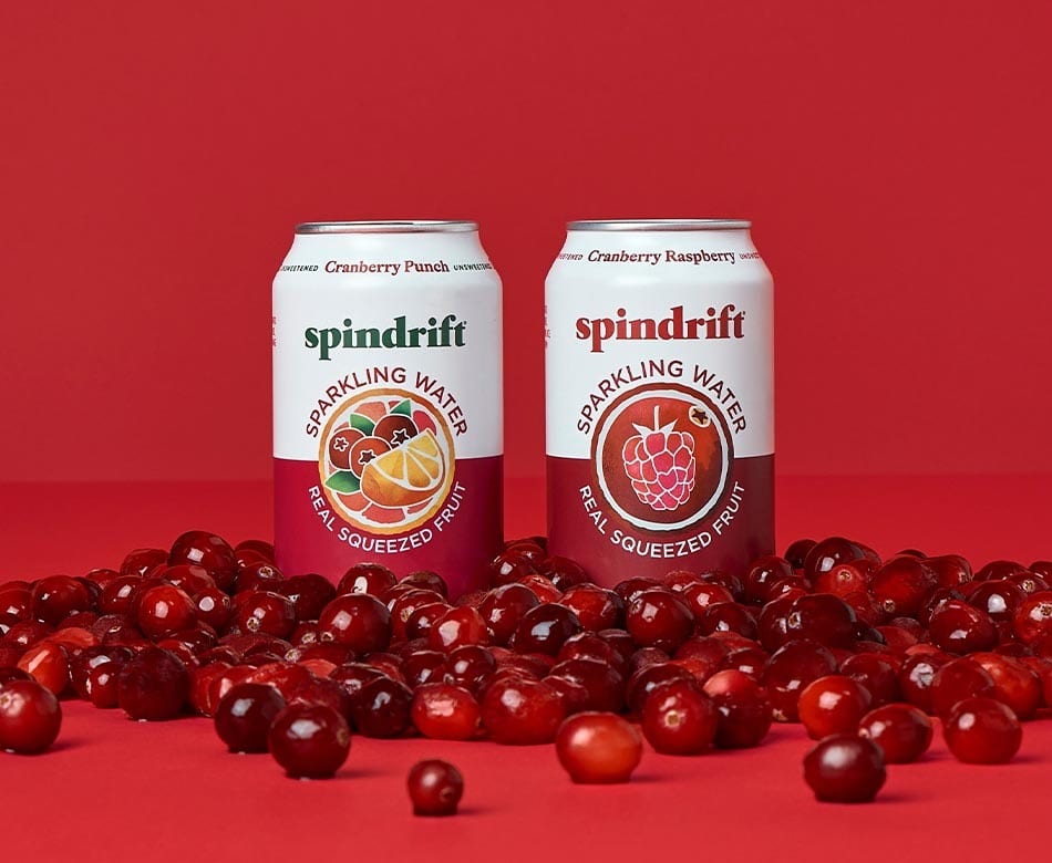 Two Spindrift sparkling water cans (Cranberry Punch, Cranberry Raspberry) amidst cranberries on red.