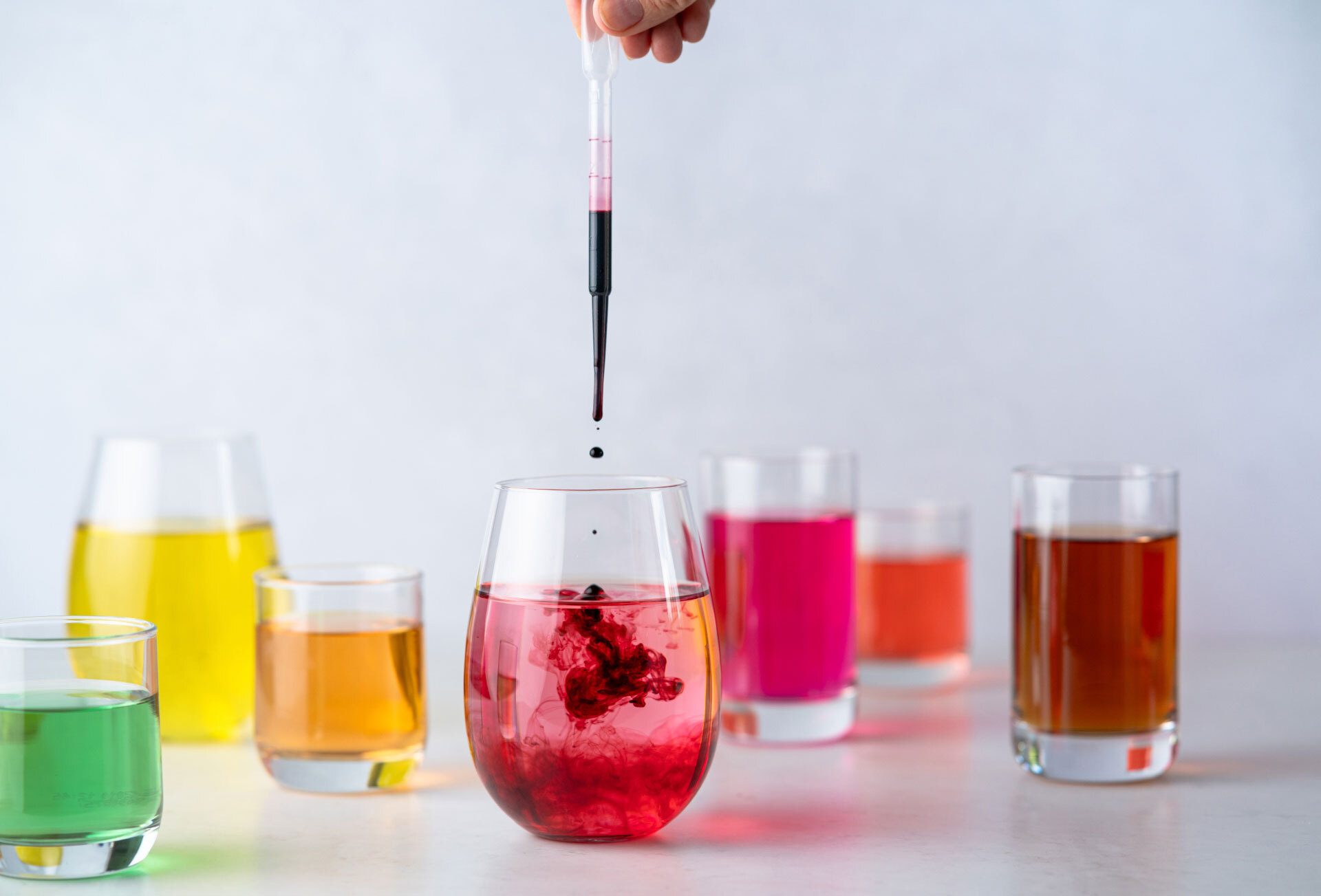 A hand drops red liquid with a dropper into a pink liquid, surrounded by other colorful glasses.