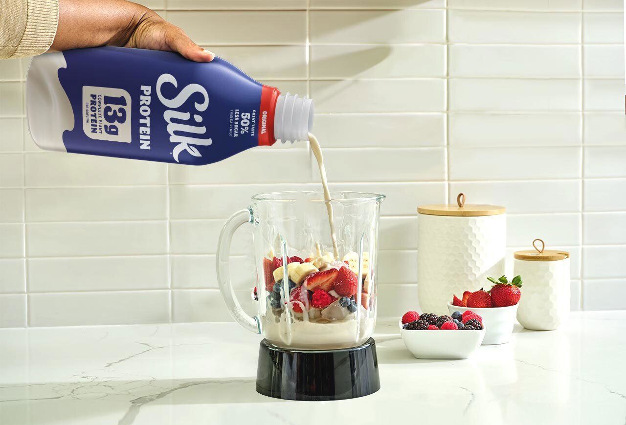 A hand pours Silk protein milk into a blender filled with berries and bananas on a kitchen counter.