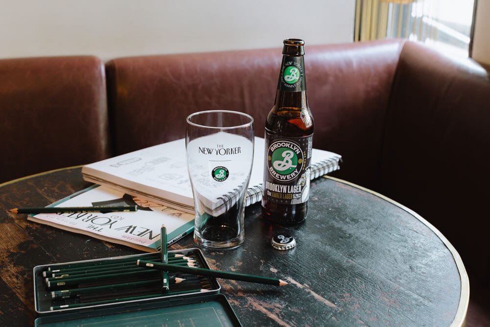 Brooklyn Brewery beer, New Yorker glass, pencils, and sketchbooks on a wooden table.