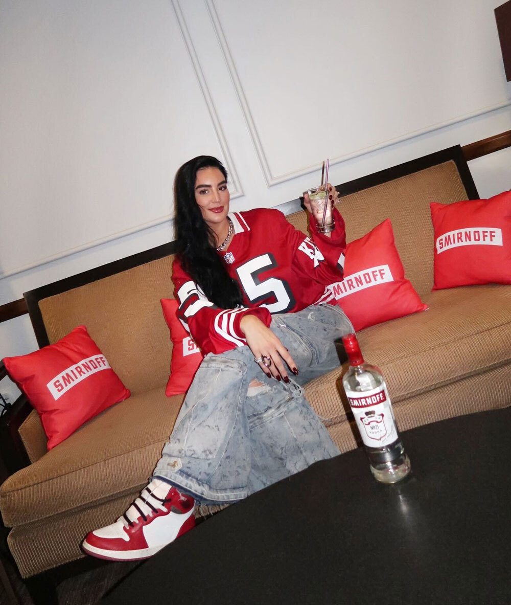 Woman in red jersey, ripped jeans, sneakers on couch with Smirnoff bottle and drink.